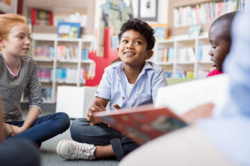 Multiethnic group of kids sitting on floor in circle around the teacher and listening a story. Discussion group of children in a library talking to woman. Smiling hispanic boy in elementary school.