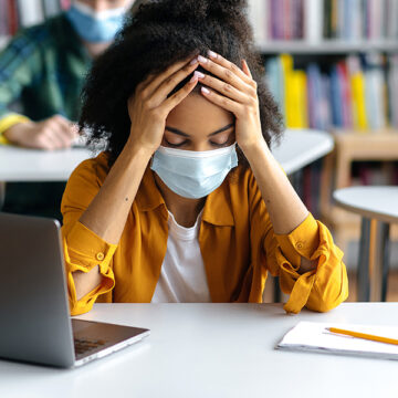 Education during pandemic. Frustrated, overworked african american student girl wearing protective medical mask sits at a table in the library, preparing for exams, tired, classmates sit at background