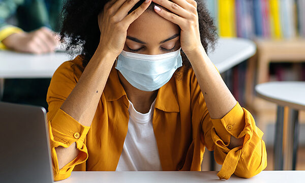 Education during pandemic. Frustrated, overworked african american student girl wearing protective medical mask sits at a table in the library, preparing for exams, tired, classmates sit at background
